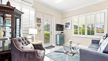 A spacious living room with a grey sofa, a glass coffee table, and a blue rug. There is a large window with blinds, a television mounted on the wall, and a decorative cabinet.