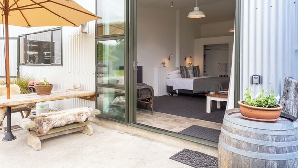 A modern, open-plan living area with a glass door leading to a bedroom, a wooden bench, a table with a fur throw, and a potted plant. - Luxury county guesthouse-continental breakfast supplied/own farm free range eggs (Omarama)