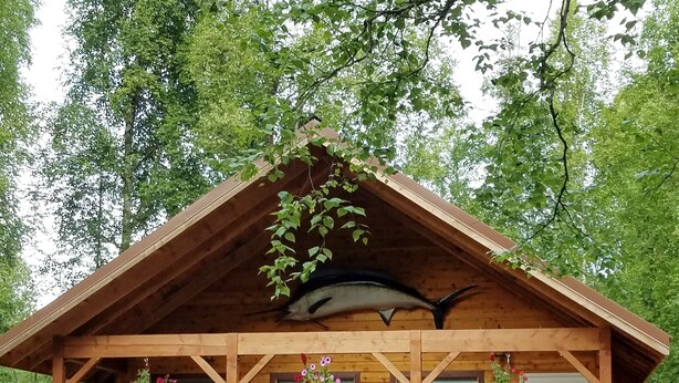 A wooden cabin with a balcony, a large fish hanging from the roof, and hanging flower baskets.