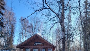 A wooden cabin with a porch, surrounded by snow-covered ground and trees.