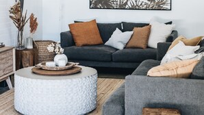 A living room with a grey sofa, a round white ottoman, and a wooden surfboard against the wall. - Cavella South Coast (Culburra Beach)