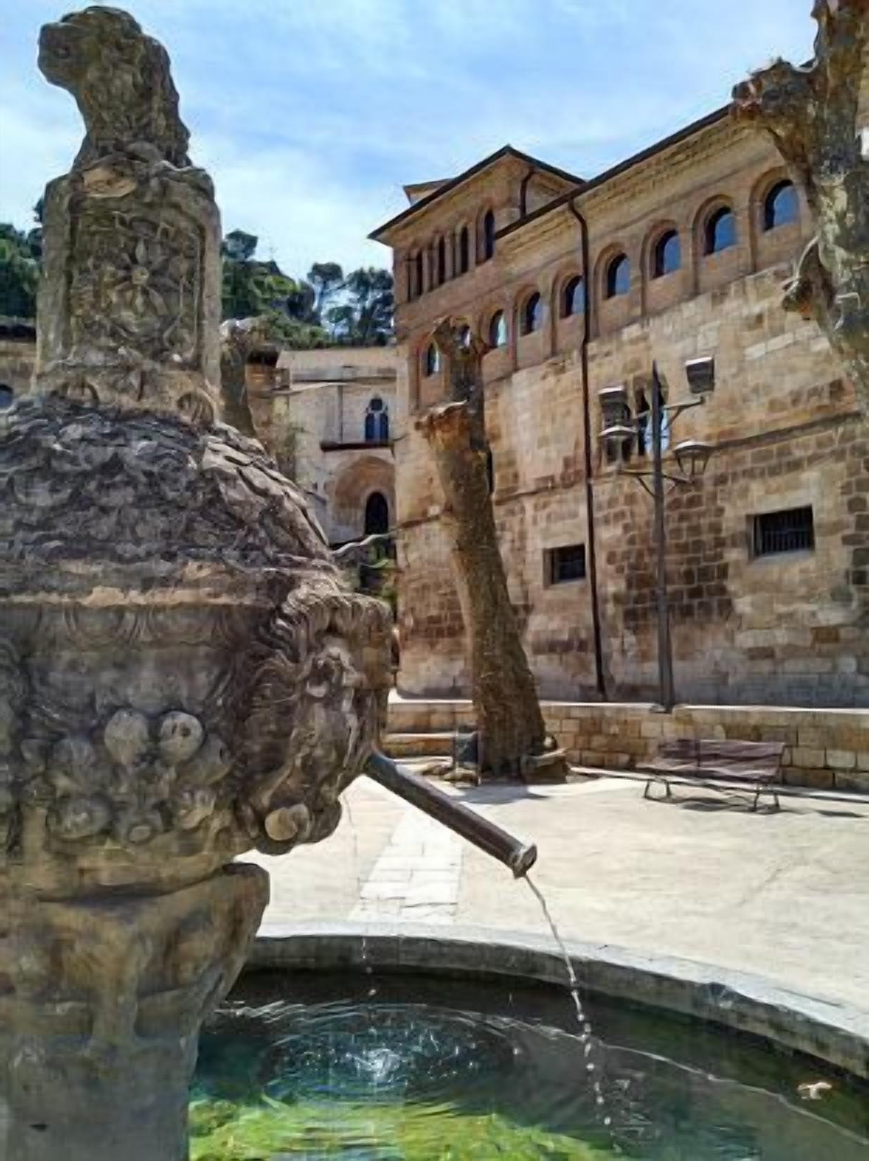 A stone fountain with water spouting from its center, surrounded by a courtyard with a building featuring arched windows and a stone wall.