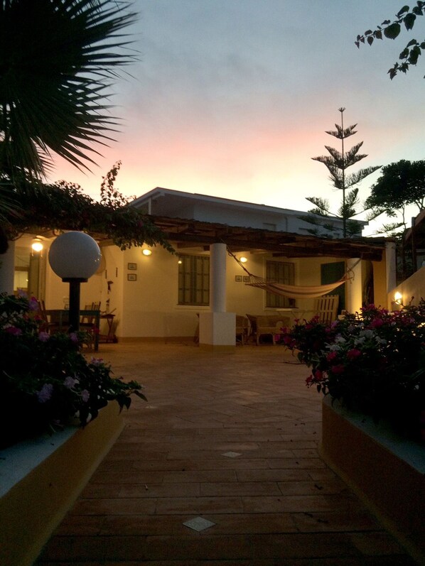 A courtyard with a wooden pathway leading to a building with a covered seating area, surrounded by plants and a hammock. - Patrizia house, independent 10 meters from the sea - San Lorenzo - Reitani - Marzamemi (noto)