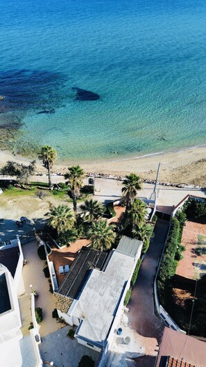 An aerial view of a coastal area with clear blue water, sandy beach, and residential buildings with flat roofs and greenery. - Patrizia house, independent 10 meters from the sea - San Lorenzo - Reitani - Marzamemi (noto)