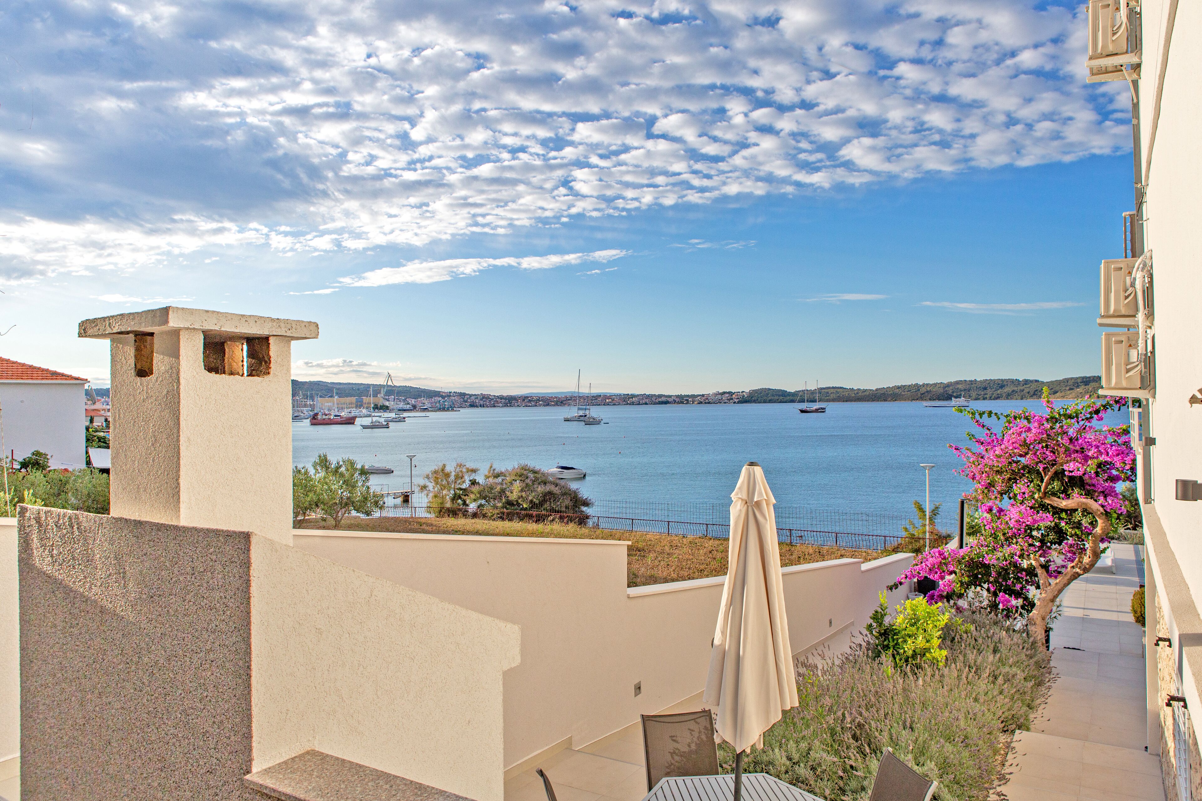 A coastal view with a balcony overlooking the sea, a small garden with bougainvillea, and a clear sky.
