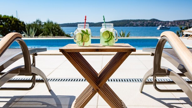 A poolside table with two mason jars of drinks, poolside chairs, and a view of the sea and hills in the background.