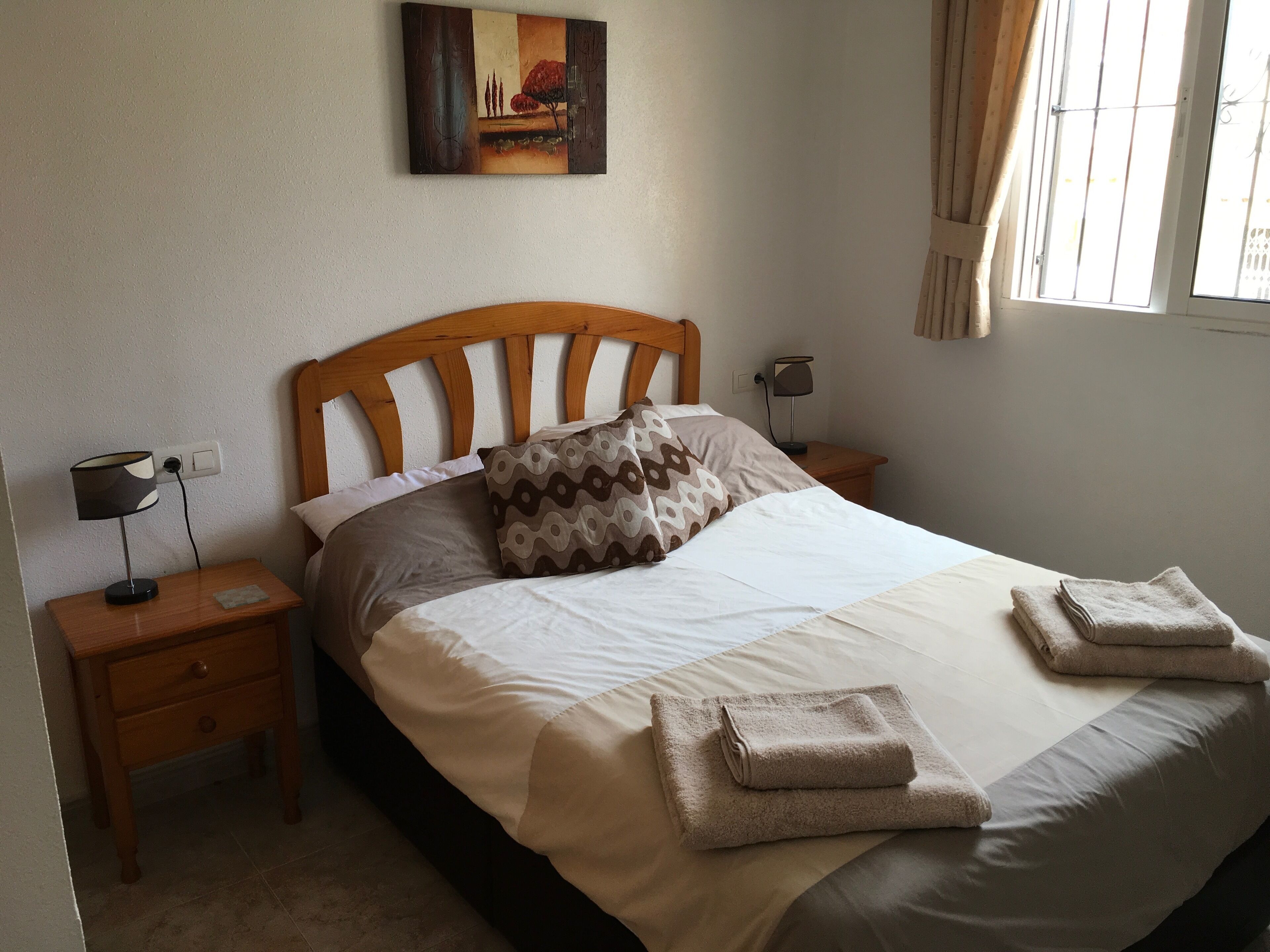 Een net opgemaakt bed met een houten hoofdbord, twee nachtkastjes met lampen, een schilderij aan de muur en een raam met gordijnen.