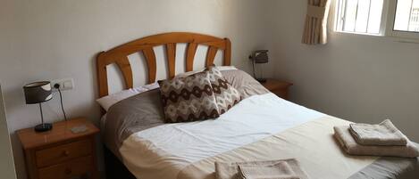 A neatly made bed with a wooden headboard, two bedside tables with lamps, a painting on the wall, and a window with curtains.