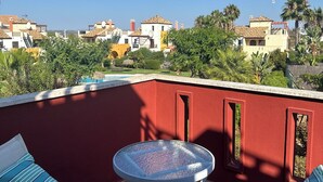 Eine Dachterrasse mit roter Wand, zwei gestreifte Loungesessel, einem kleinen runden Tisch und Blick auf ein Wohngebiet mit Palmen und Häusern.