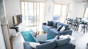 Ein modernes Wohnzimmer mit einer grauen Sofagarnitur, einem Flachbildfernseher und einem Essbereich mit Tisch und Stühlen.