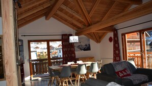Ein geräumiger Wohnbereich mit Esstisch, Stühlen und einer Couch. Durch die Fenster ist ein Balkon mit Bergblick zu sehen.