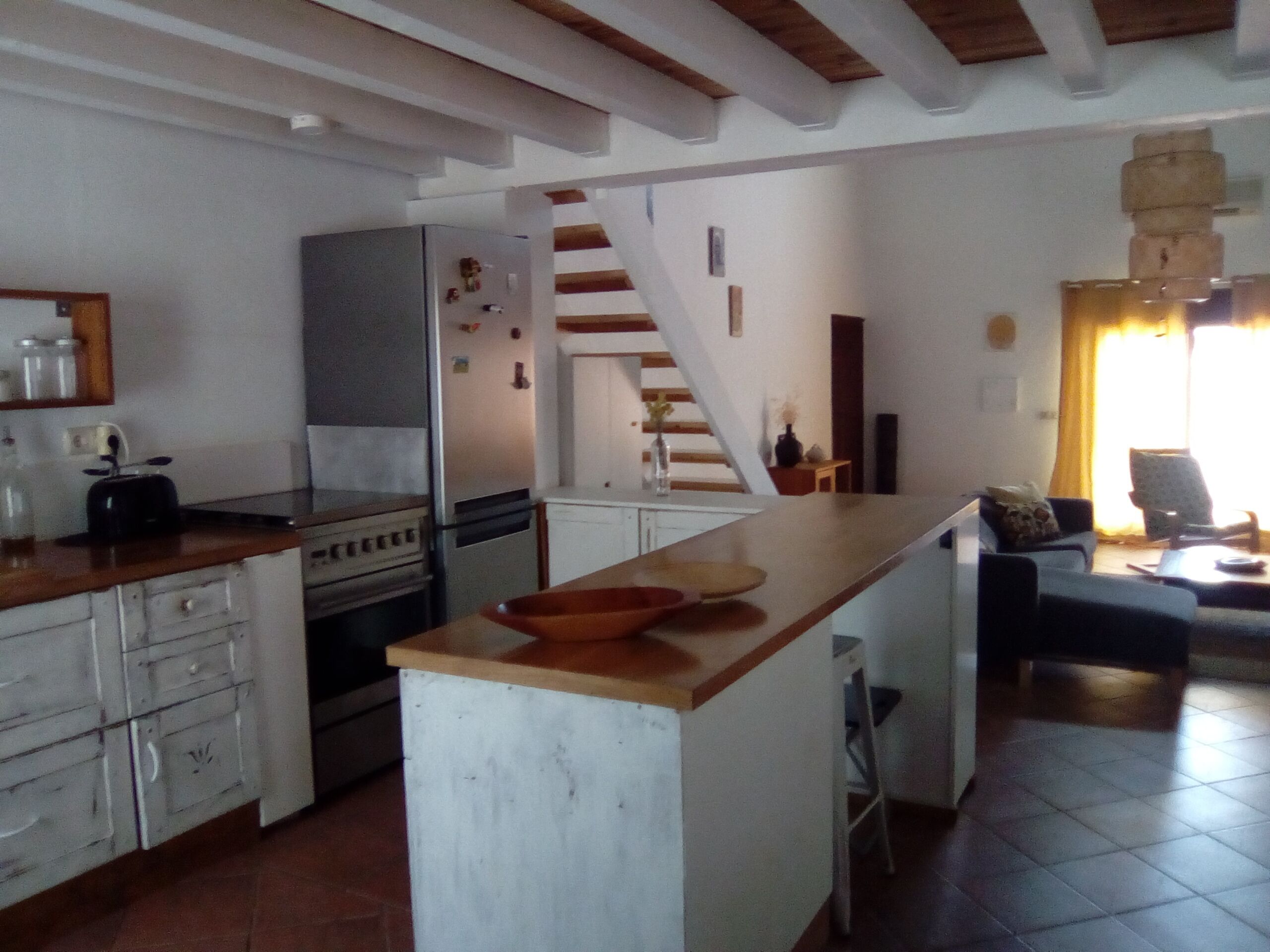 A compact kitchen with a central island, a built-in oven, and a refrigerator. A staircase leads to a living area with a sofa and a chair.