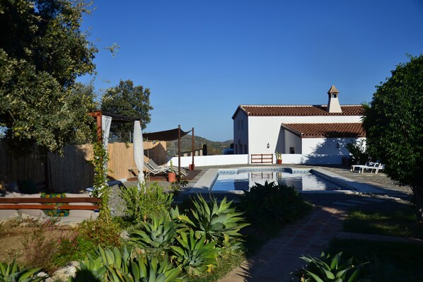 A white building with a tiled roof and a swimming pool. There are plants and a wooden fence in the foreground. - The African House, rural getaway between Ronda and Málaga (Tolox)