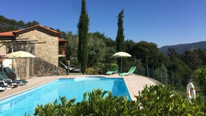 Piscina con sillones de playa y sombrillas, rodeada de vegetación y un edificio de piedra.