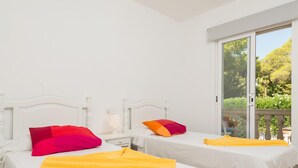 Une chambre d’hôtel avec deux lits, chacun recouvert d’une courtepointe blanche et agrémenté de coussins colorés. Une fenêtre offre une vue sur la verdure environnante.