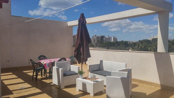 A balcony with wicker furniture, a table with a pink tablecloth, and a view of buildings in the distance.