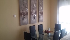 A dining area with a glass table and six chairs, three abstract paintings on the wall, and a window with curtains. - Beautiful duplex apartment with pool 200 meters from the beach and golf (el campello muchavista)