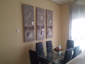 Ein Essbereich mit einem Glastisch und sechs Stühlen, drei abstrakte Gemälde an der Wand und ein Fenster mit Vorhängen.