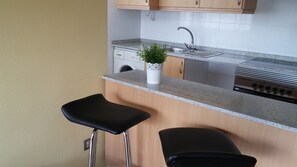 Eine Küche mit einer Granitarbeitsplatte, einem Edelstahlspülbecken und einem eingebauten Backofen. Auf der Arbeitsplatte befinden sich zwei schwarze Barhocker und ein Blumentopf.
