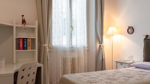 A bedroom with a bed, a desk with books, a chair, a lamp, and a window with curtains.