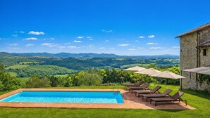 A swimming pool with lounge chairs and umbrellas, surrounded by a stone building and a view of rolling hills.