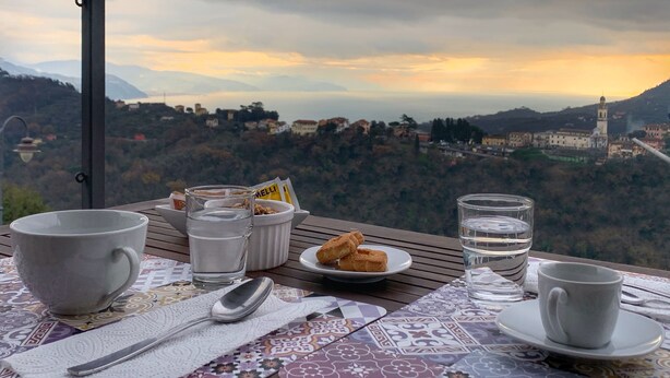 Ein Tisch mit Tasse, Glas Wasser, Teller mit Keksen und Löffel, mit Blick auf eine malerische Stadtlandschaft mit Bergen und einer Kirche.