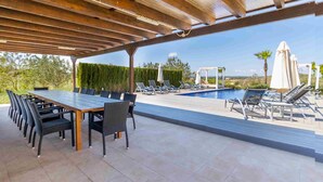 An outdoor patio with a long dining table and chairs, a pool area with lounge chairs and umbrellas, and a view of the landscape.