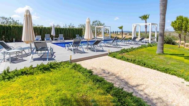 A pool area with lounge chairs and umbrellas, surrounded by greenery and a clear sky.