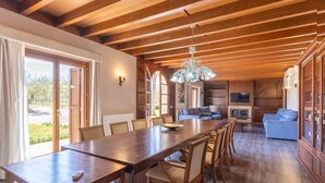 A spacious dining area with a long wooden table and chairs, a chandelier, and a view of the outdoors through large windows.