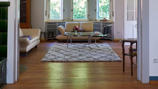Ruime kamer met houten vloer, een bank, een eettafel en een raam met uitzicht op groen.