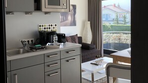 A modern kitchen with grey cabinets, a microwave, and a coffee machine. There is a sofa and a table with a chair in the living area, and a balcony with a view of houses and trees. - City-Apparte One - Modern apartment building in a central location (Gronau)