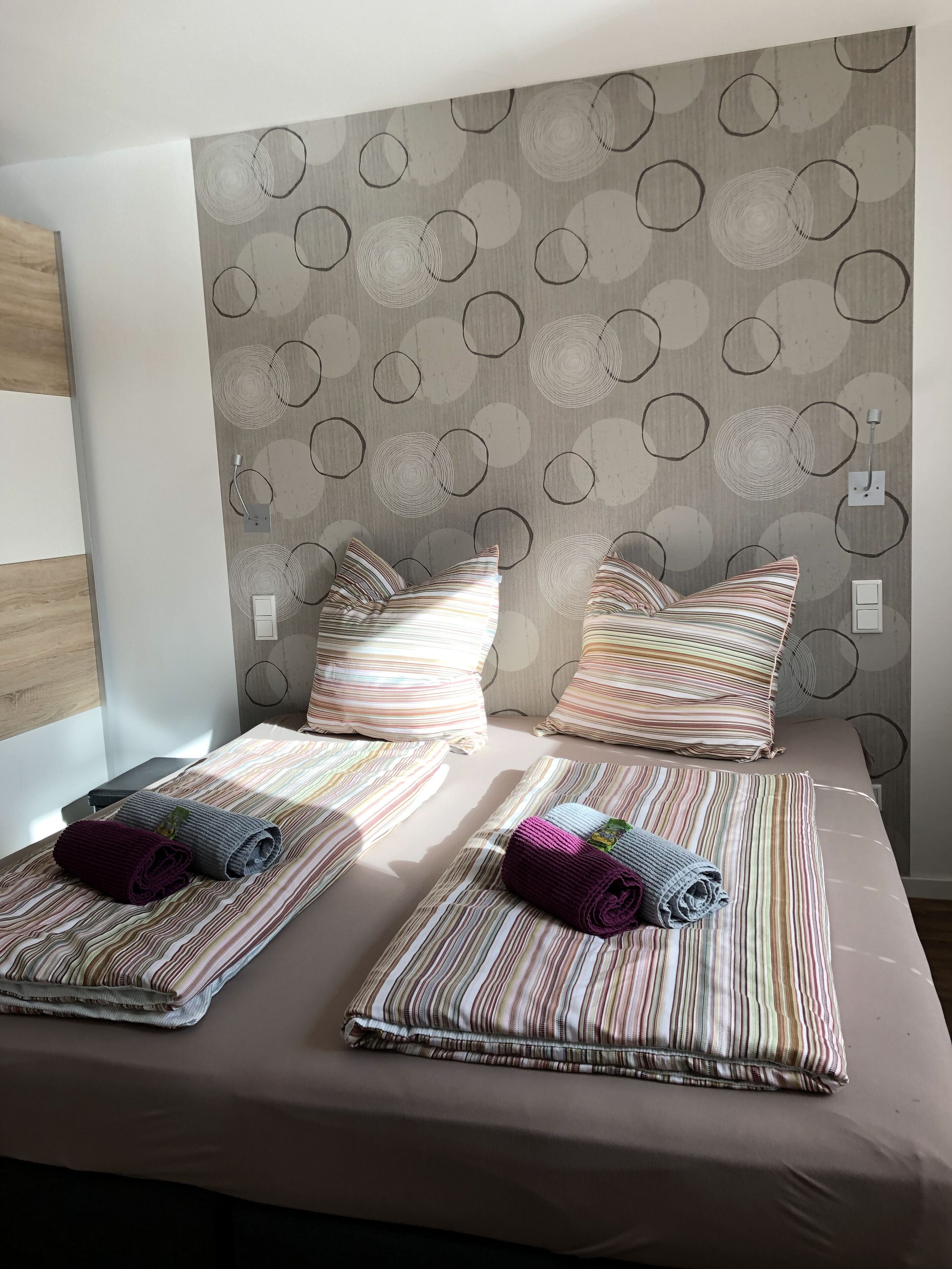 A bed with striped bedding and two pillows, a wall with circular patterns, and a wooden panel on the left.