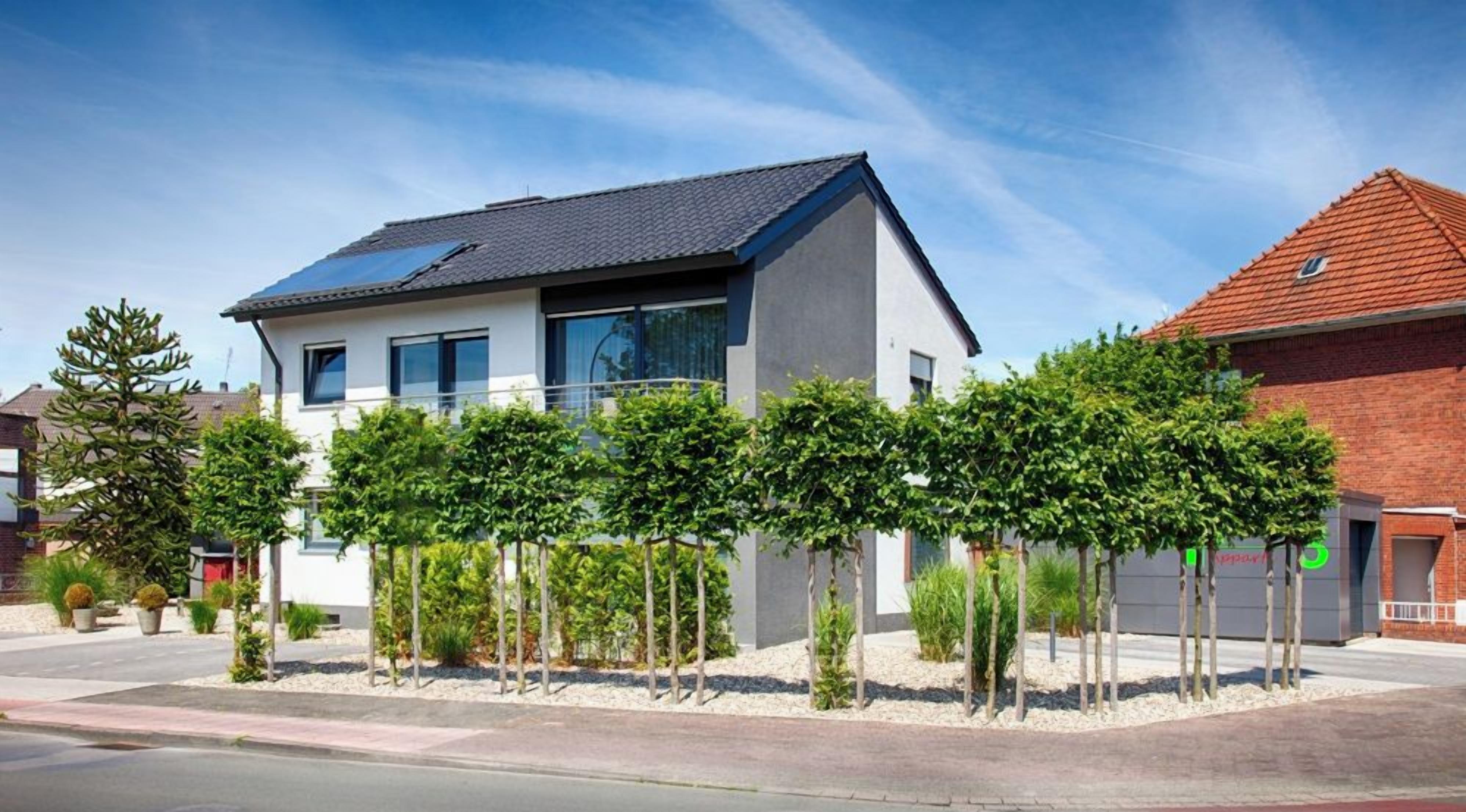Une maison moderne à deux étages, dotée d’un toit plat et de panneaux solaires, entourée d’un jardin bien entretenu où poussent de jeunes arbres et arbustes.