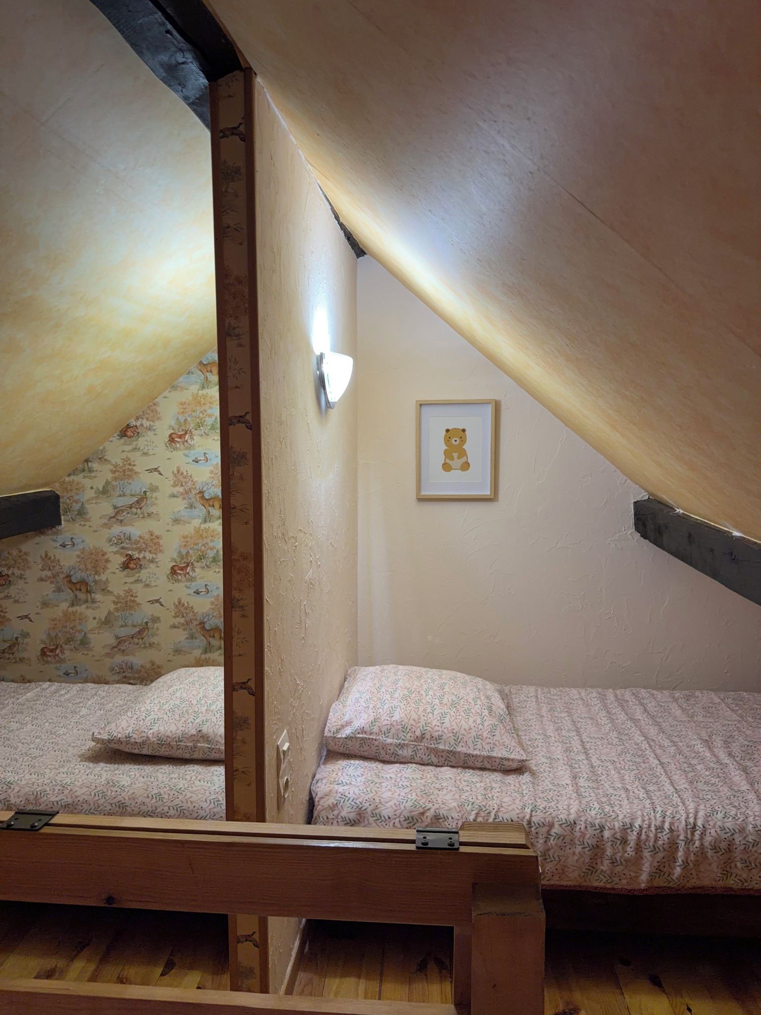 A small, single-bed room with a sloped ceiling, a wooden beam structure, and a framed picture on the wall.