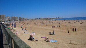 Una playa concurrida, gente tomando el sol y jugando, cielo azul despejado y edificios al fondo.