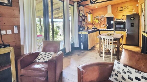 Un acogedor interior de cabaña con techo de madera, un comedor con mesa y sillas, una cocina con refrigerador y estufa, y un ventanal con cortinas.