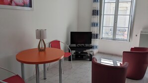 A small living area with a round orange table, two red chairs, a TV, and a window with patterned curtains.