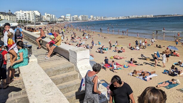 A crowded beach with people sunbathing and swimming, a pier in the background, and buildings along the coastline.