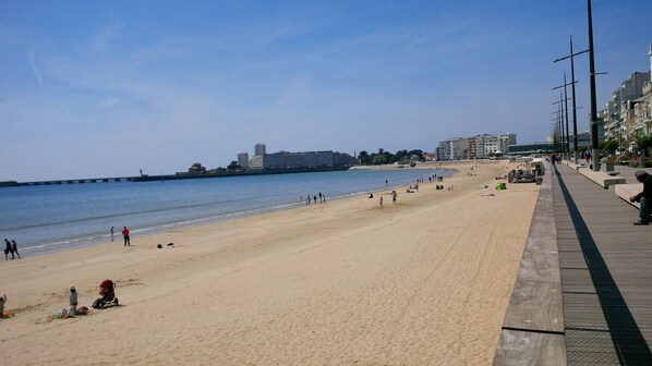 A wide sandy beach with a promenade on the right, a calm sea, and buildings in the background.