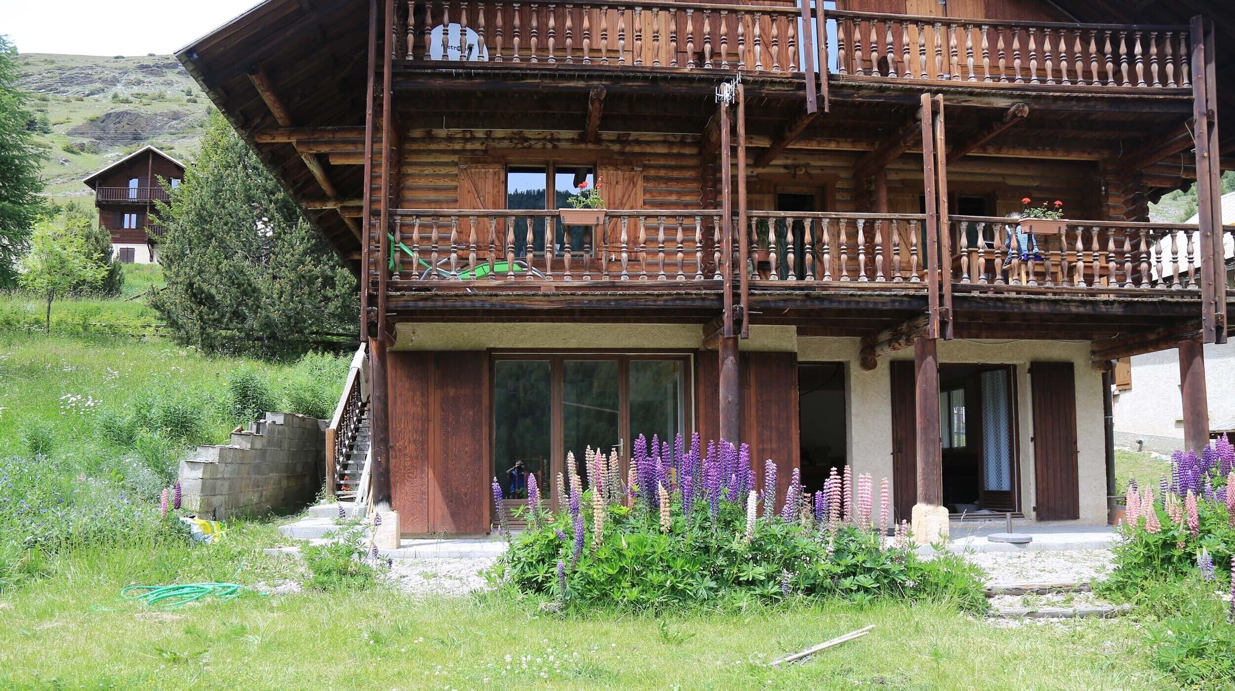 A wooden chalet with a balcony, large windows, and a garden with purple flowers.