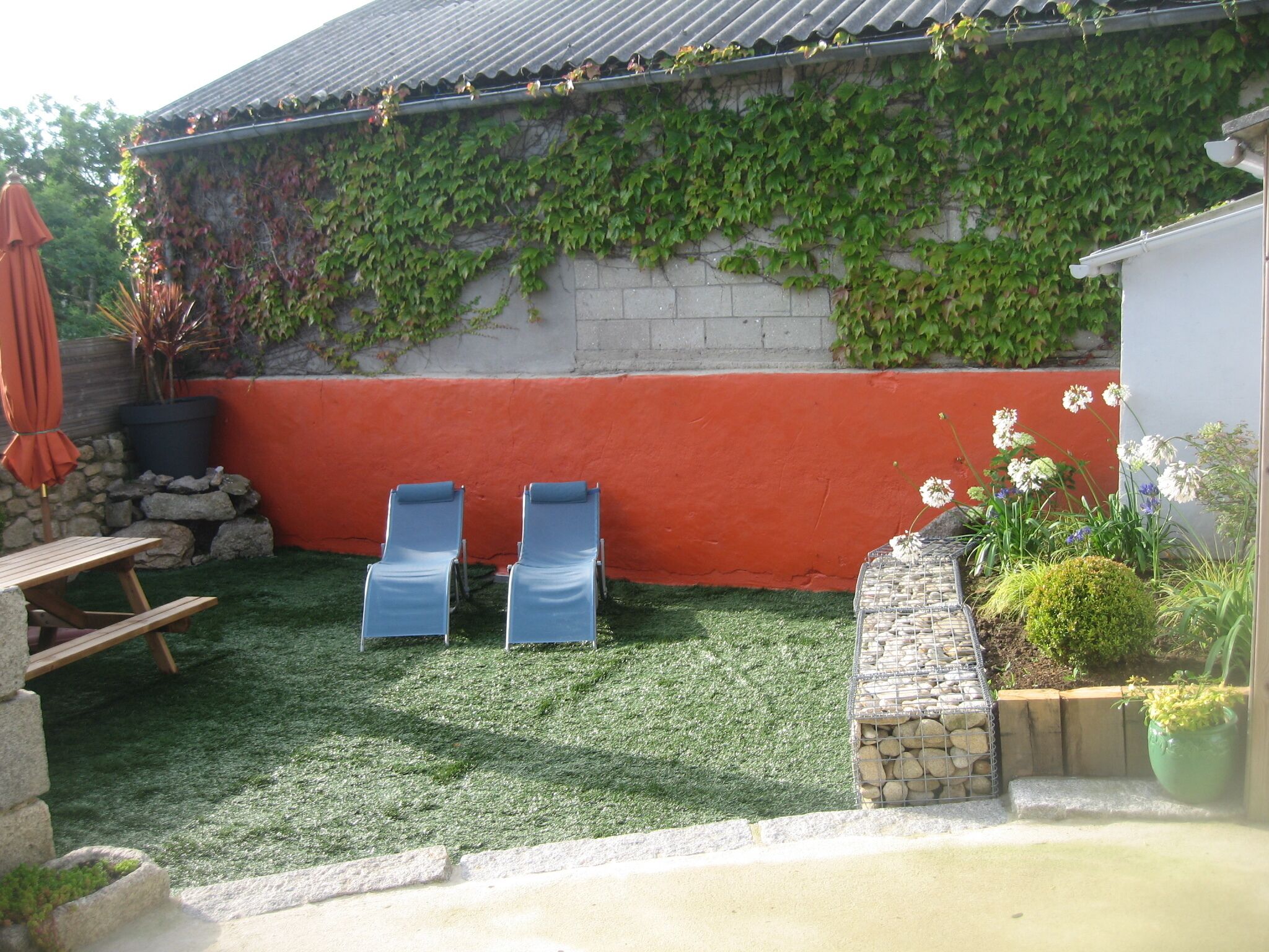 Een achtertuin met kunstgras, twee blauwe loungestoelen, een stenen muur en een potplant.