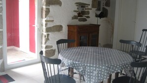 A dining area with a round table covered by a polka dot tablecloth, surrounded by six black chairs. A wooden cabinet is against the wall, and a door with a red frame is visible in the background.