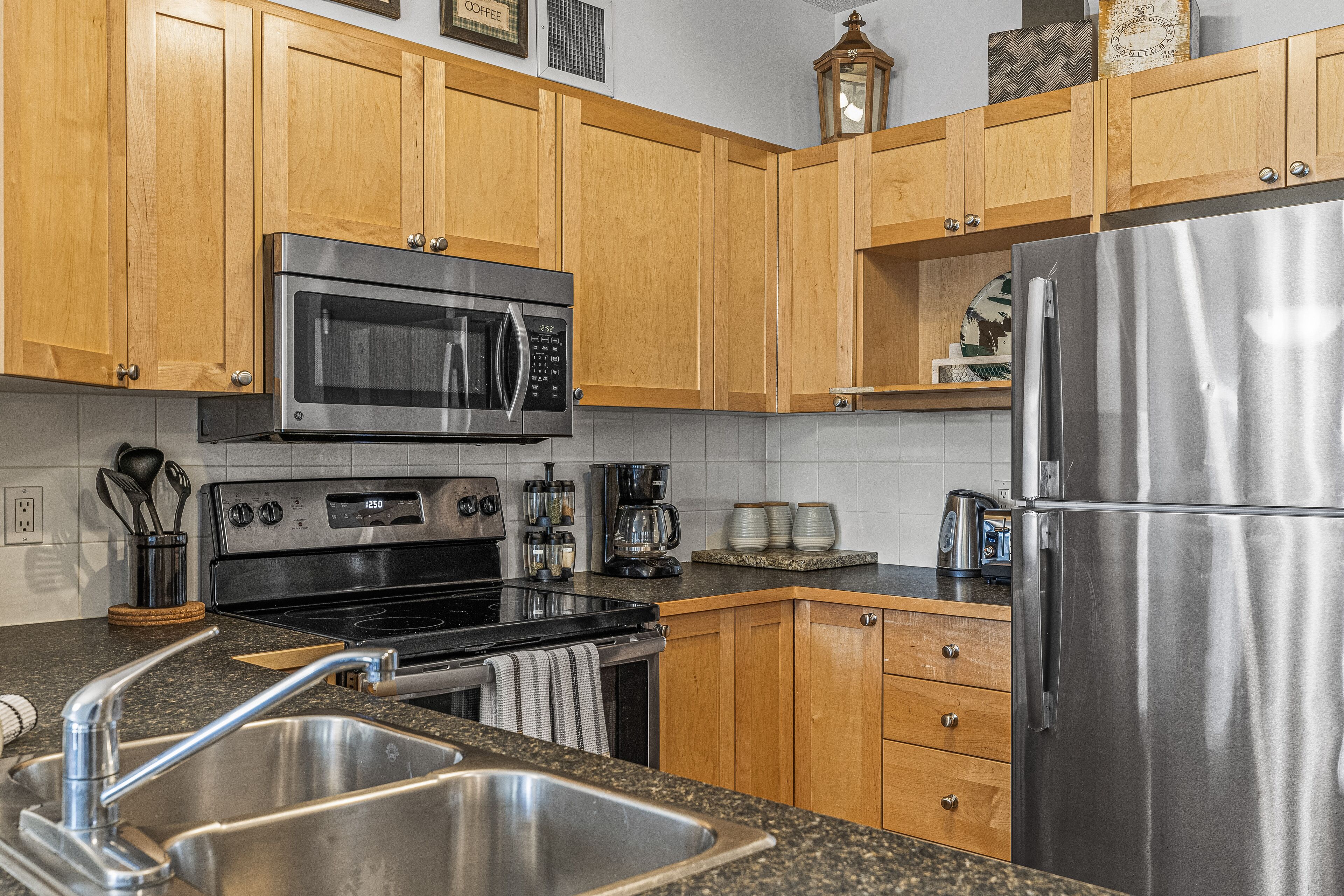 A modern kitchen with wooden cabinets, stainless steel appliances including a microwave, oven, and refrigerator, and a granite countertop.