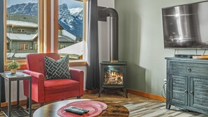 Ein gemütliches Wohnzimmer mit einem roten Sessel, einem Kamin, einem Fernseher und Blick auf schneebedeckte Berge durch große Fenster.