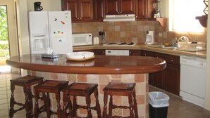 Eine Küche mit Holzschränken, einem weißen Kühlschrank, einer Mikrowelle und einer Kaffeemaschine. Vier Holzbarhocker stehen an der Kücheninsel.