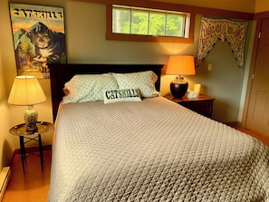 A neatly made bed with a quilted cover, a lamp, a framed poster on the wall, and a window with a view of trees.