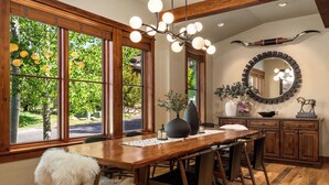 Un comedor con mesa de madera, sillas, una araña y ventanas amplias con vistas a la vegetación.