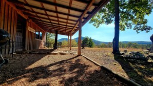Un espacio al aire libre cubierto con piso de grava, mesas y sillas de madera. Se ve una parrilla a la izquierda. El área está rodeada de árboles y una vista de colinas distantes.