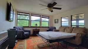 Una habitación con cama, dos sillones, un radiador, un ventilador de techo y ventanas amplias con vistas al verdor.