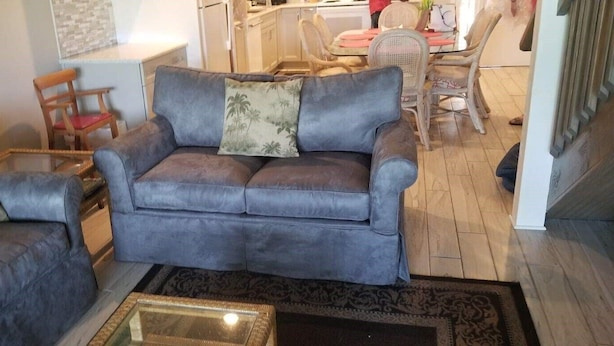 Ein Wohnzimmer mit einer grauen Couch, einem passenden Sessel und einem kleinen Couchtisch. Im Hintergrund ist eine Essecke mit Tisch und Stühlen zu sehen.